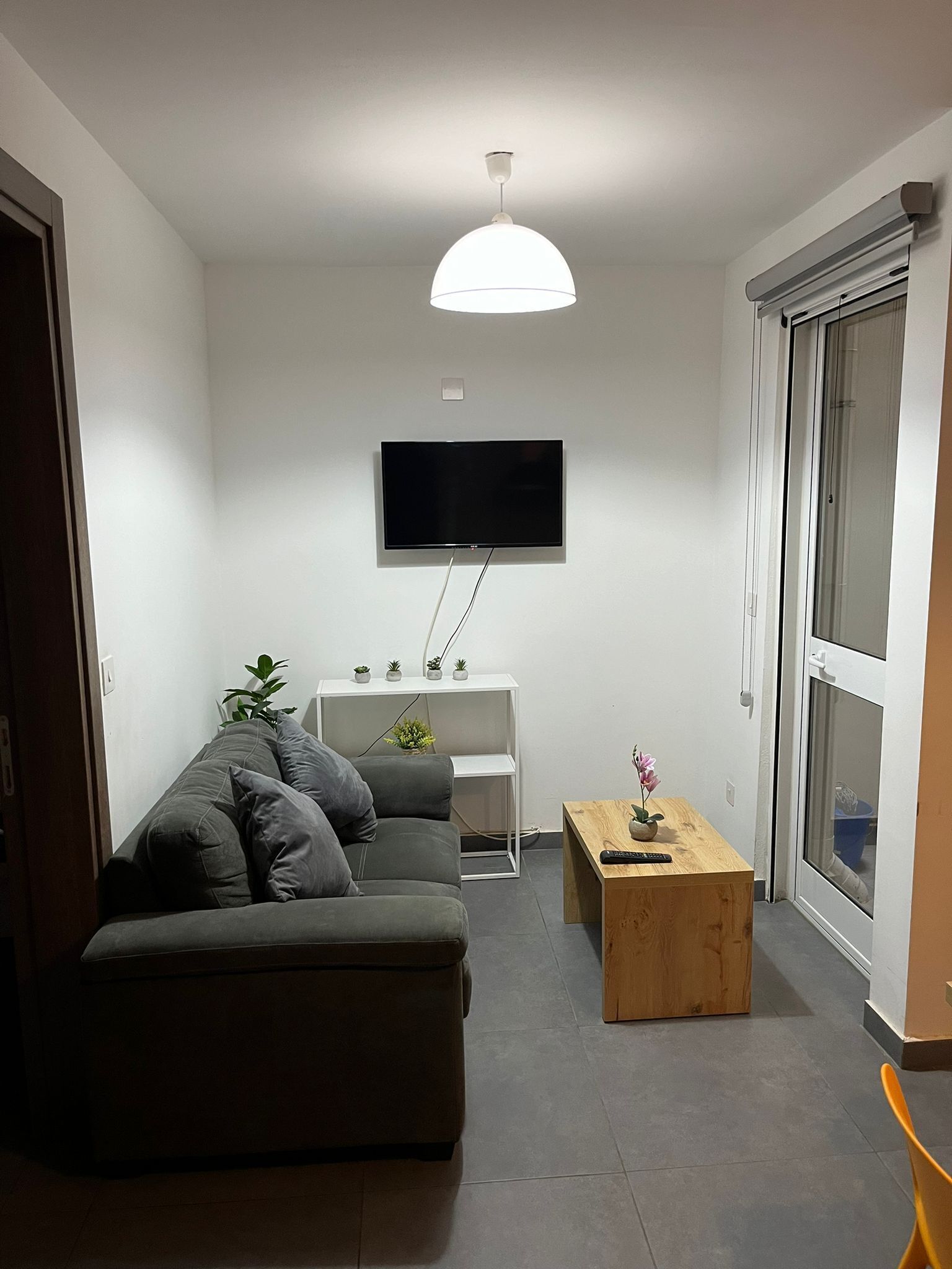 A modern living room with a grey sofa, a wooden coffee table, a TV mounted on the wall, and a sliding glass door to the right.