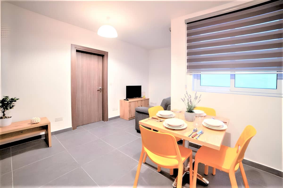 A modern dining area with a wooden table set for four, surrounded by orange chairs. A television is placed on a wooden cabinet against the wall.