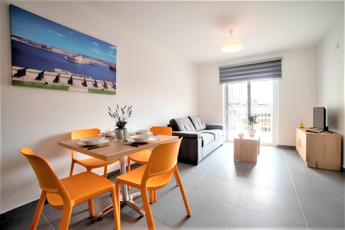 A modern living room with a dining area, a grey sofa, a wooden cabinet, and a large window with blinds.