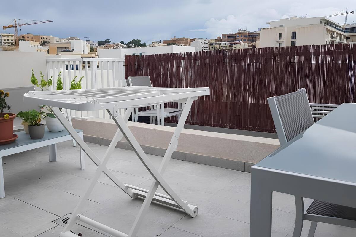 A rooftop terrace with a white table and chairs, potted plants, and a view of the cityscape.