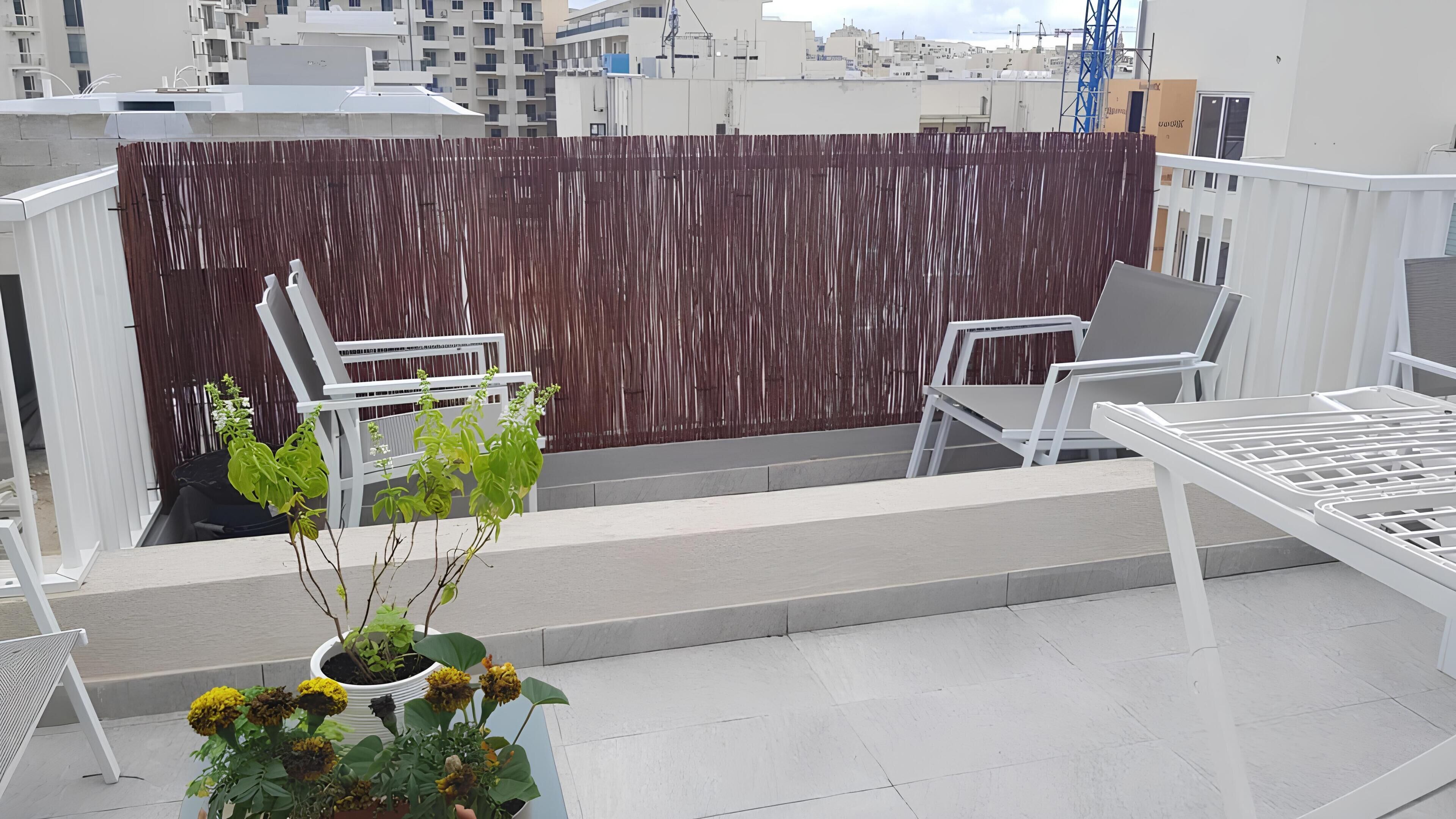 A rooftop terrace with white metal furniture, a bamboo fence, and potted plants.