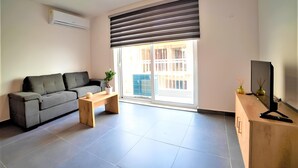 A modern living room with a grey sofa, a wooden coffee table, a TV on a wooden stand, and a large window with blinds. - Elise Apartments (Gzira)