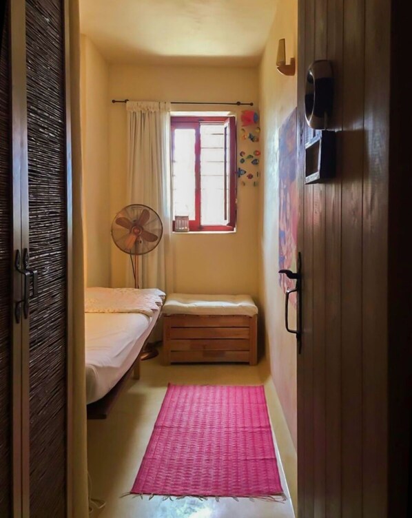 Ein schmales Zimmer mit einem Bett, einer kleinen Bank, einem Ventilator und einem Fenster mit Vorhängen.