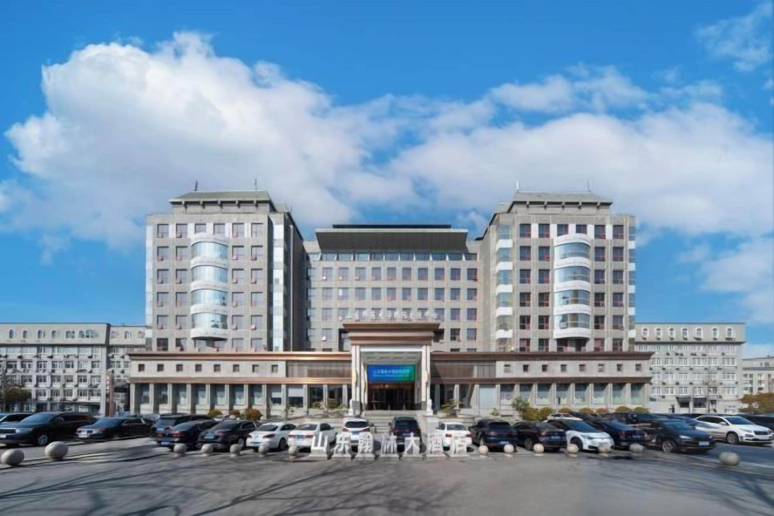 A large building with multiple floors, a central entrance, and a parking lot with several cars.