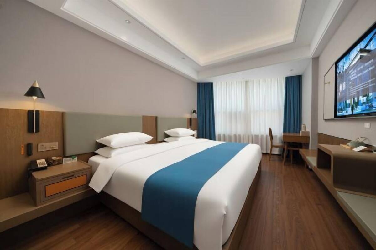 A hotel room with a large bed, bedside tables, a desk, a chair, a TV, and a window with curtains.