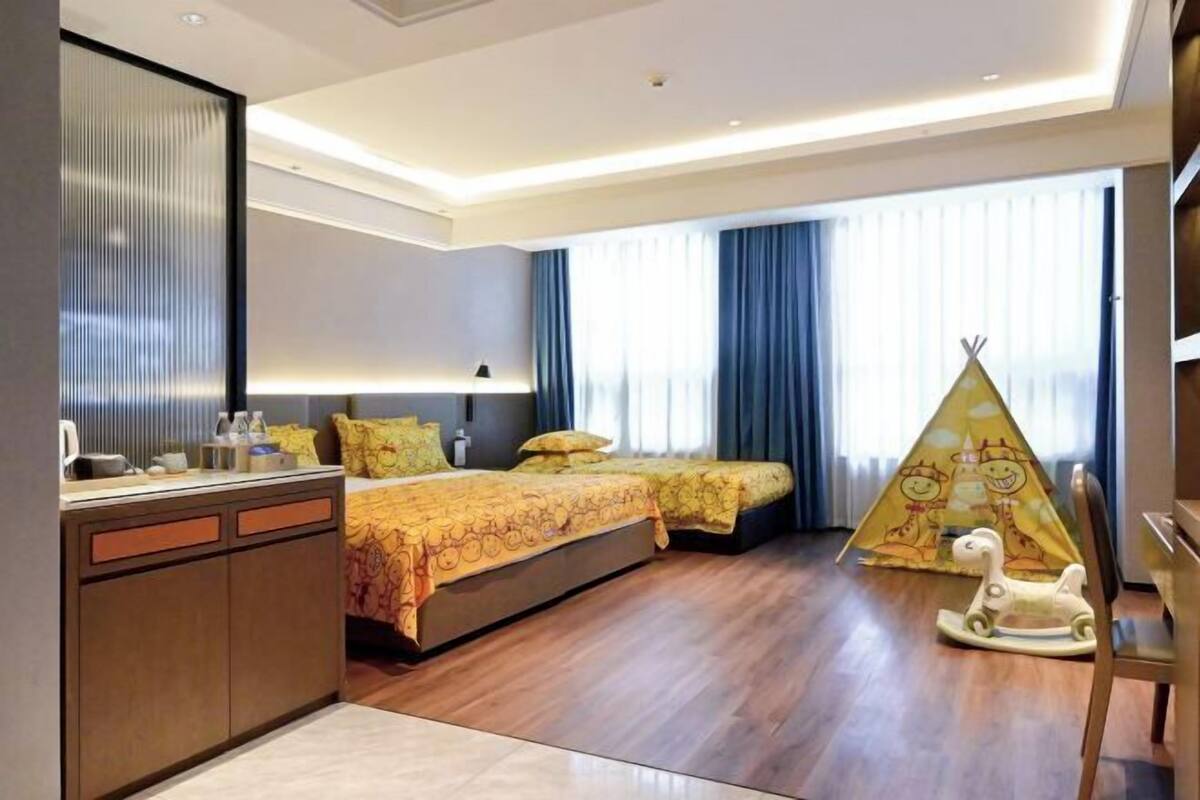 A hotel room with a bed, a desk, a chair, a window with curtains, and a play tent.