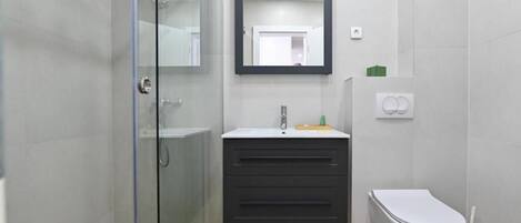 Un baño moderno con cabina de ducha de vidrio, tocador oscuro con espejo y un inodoro blanco.