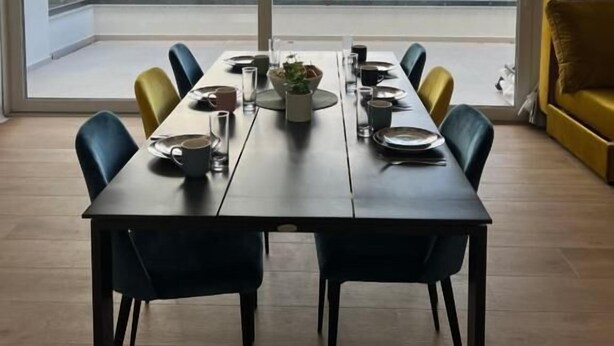 A dining area with a table set for six, large windows offering a view of the sea and cityscape, and a yellow armchair to the side.