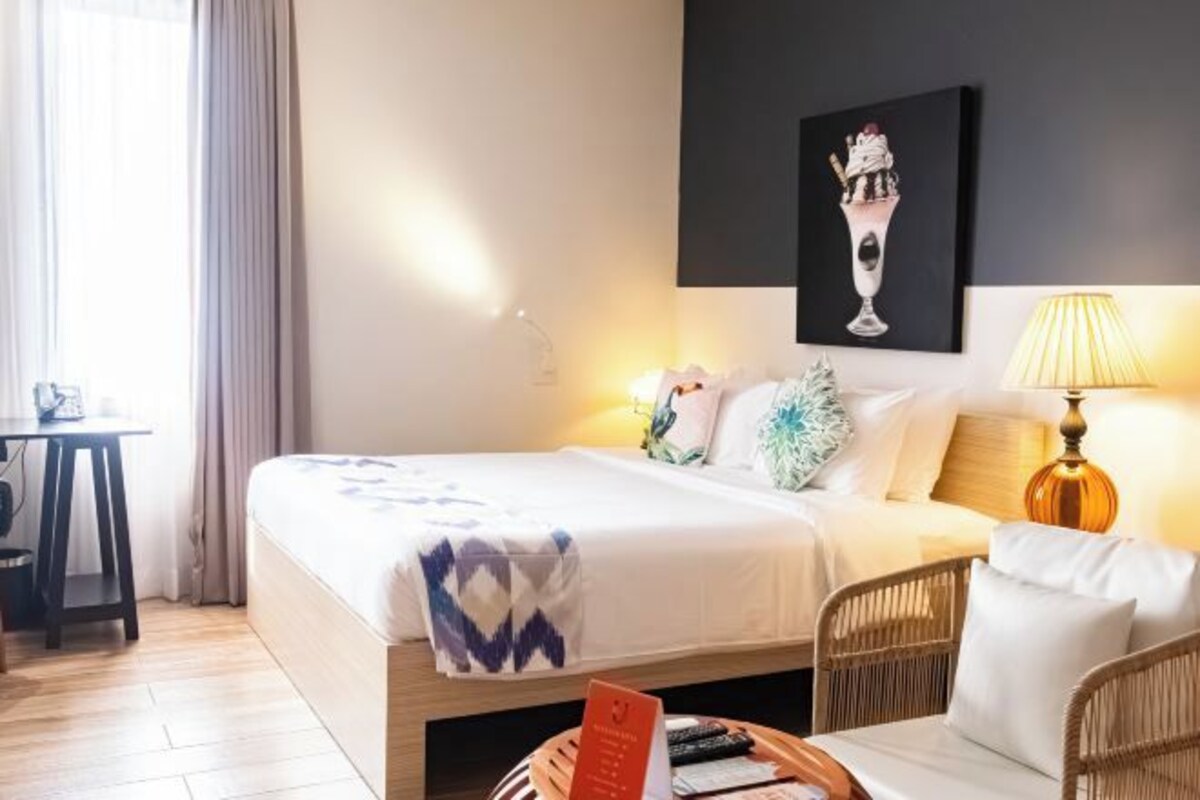 A hotel room with a bed, a desk, a chair, a lamp, and a painting on the wall.