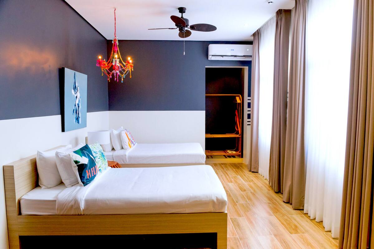 A hotel room with two beds, a chandelier, ceiling fan, air conditioning unit, and a view of the outside.