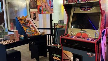 Una sala de juegos con una máquina de NBA JAM, un juego de ajedrez y coloridas obras de arte en las paredes.