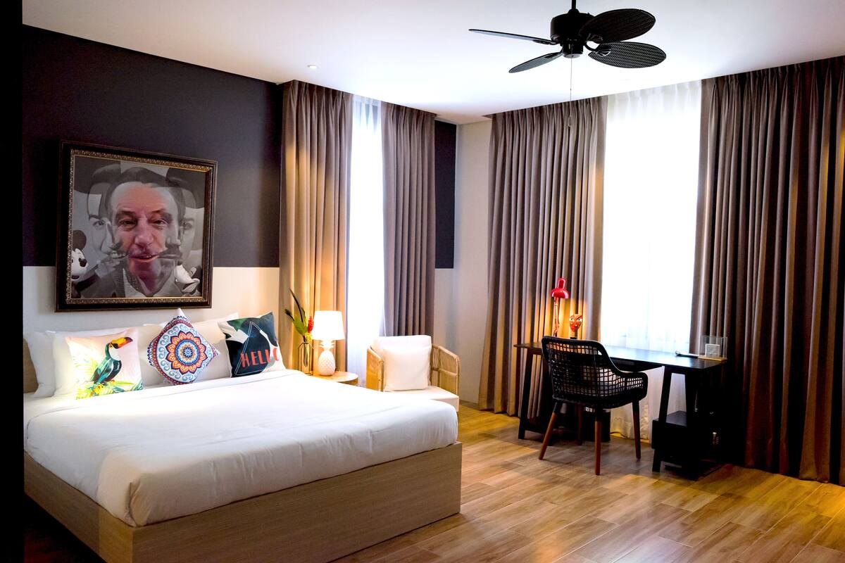 A hotel room with a bed, a desk, a chair, a ceiling fan, and curtains.