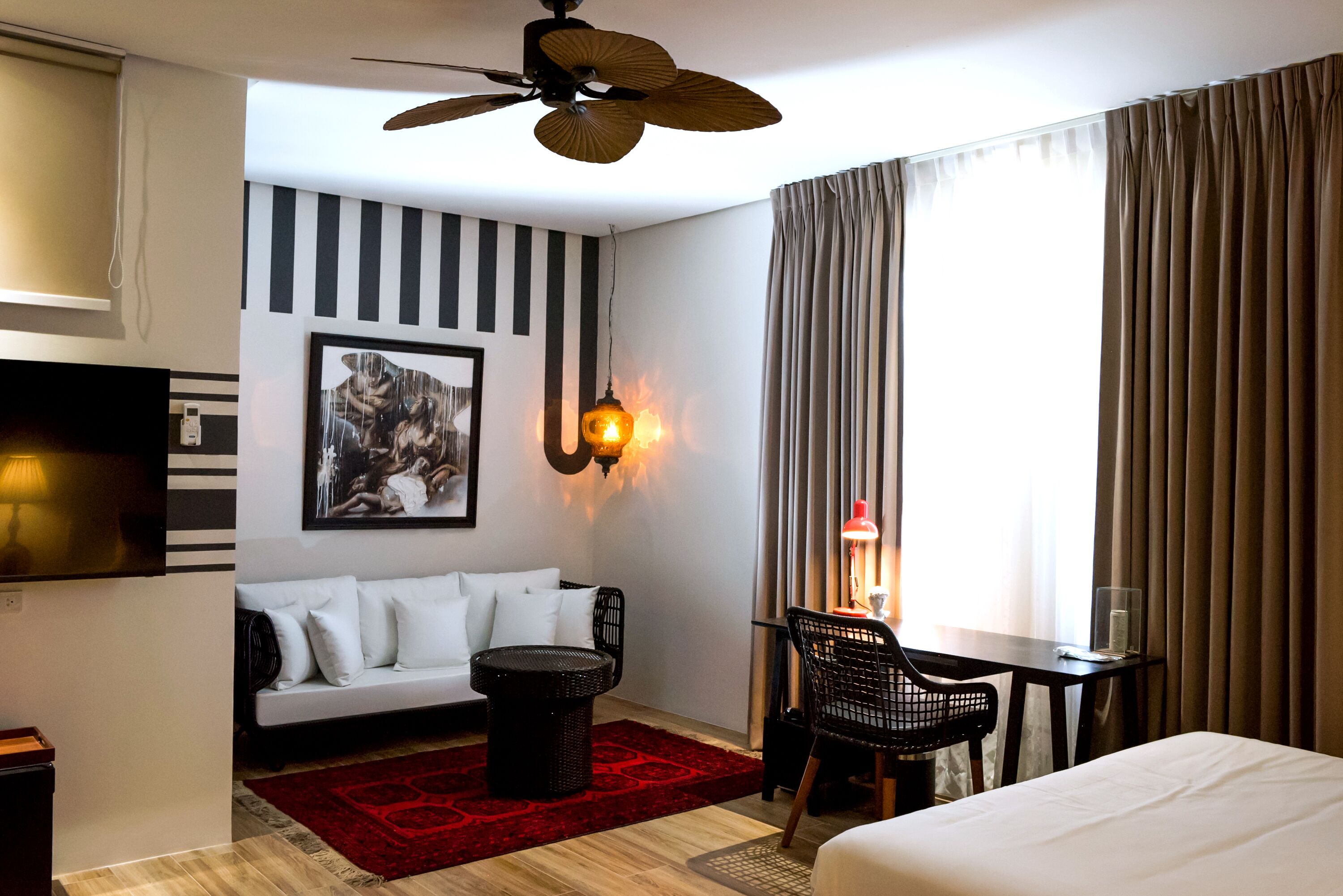 Ein Hotelzimmer mit Bett, Schreibtisch mit Stuhl, Sofa, einem roten Teppich, Deckenventilator, Wand-Fernseher und einem Gemälde.