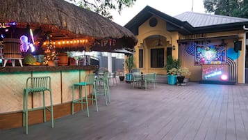 Zona de bar al aire libre con techo de paja, sillas verdes y un letrero de neón.