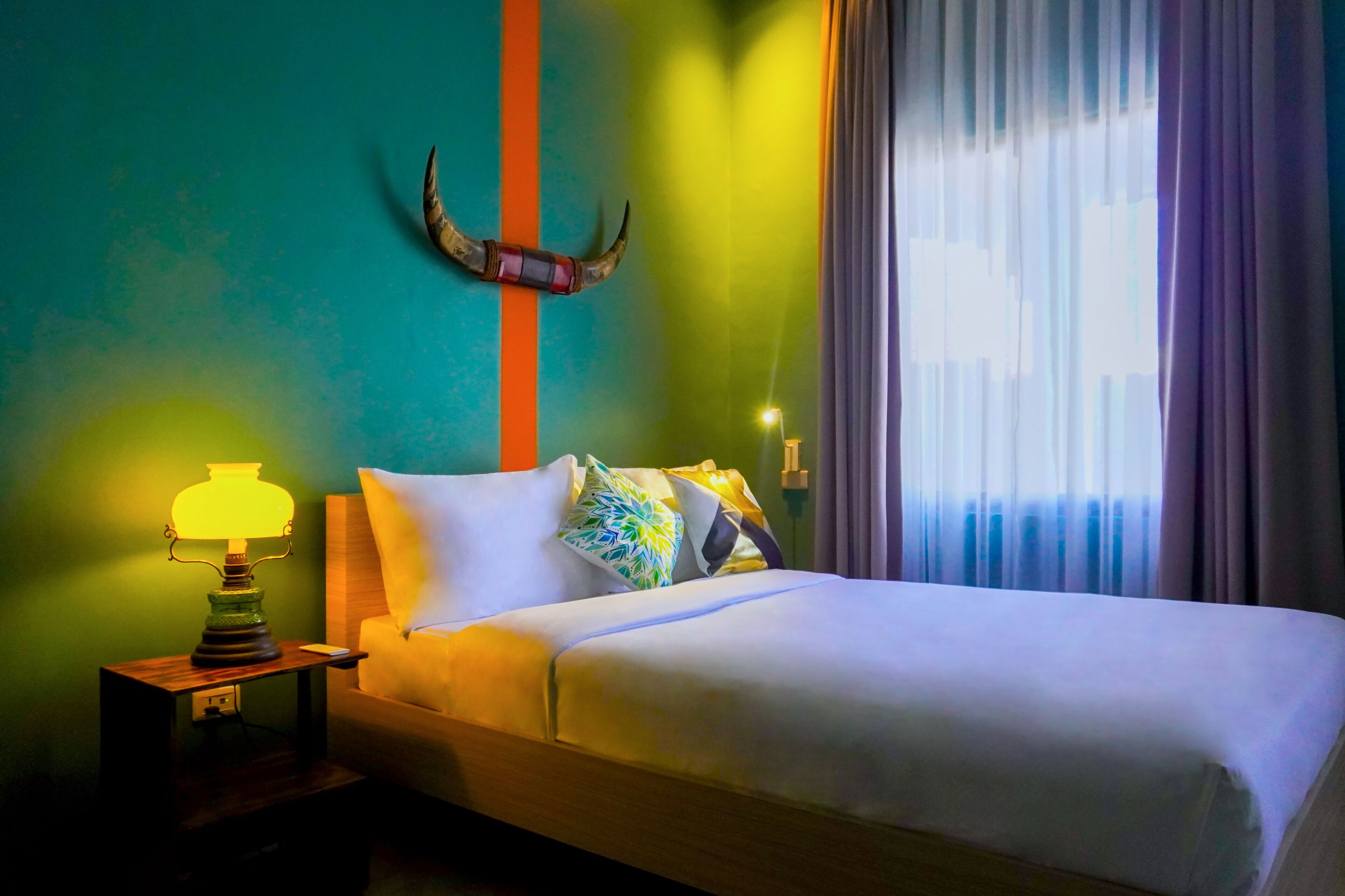 Uma cama com roupa de cama branca, uma luminária sobre uma mesa de cabeceira de madeira, um chifre decorativo na parede e uma janela com cortinas.
