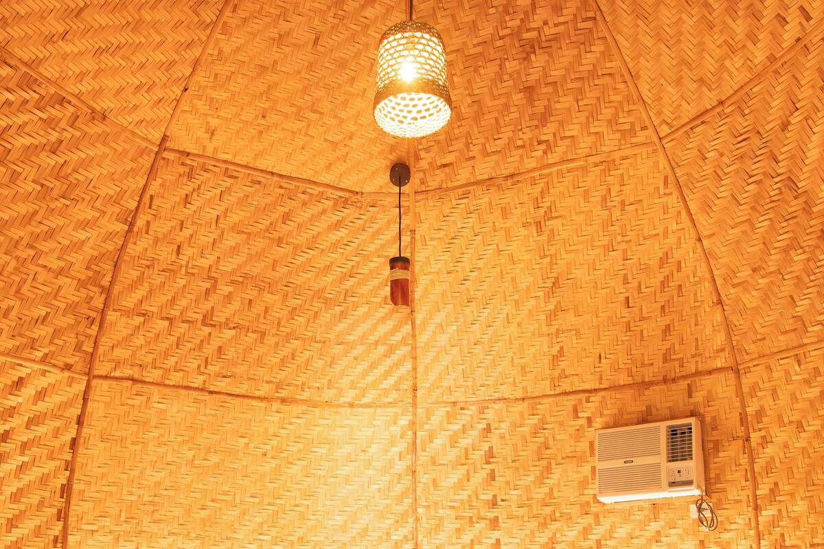 A room with a woven ceiling, a bed with white bedding, two bedside lamps, and a wall-mounted air conditioner.
