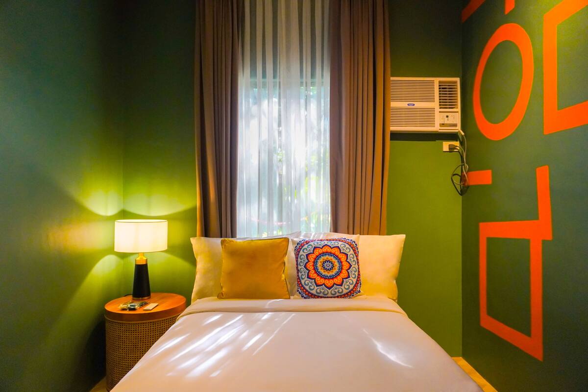 A hotel room with a bed, a nightstand with a lamp, a window with curtains, and an air conditioning unit on the wall.