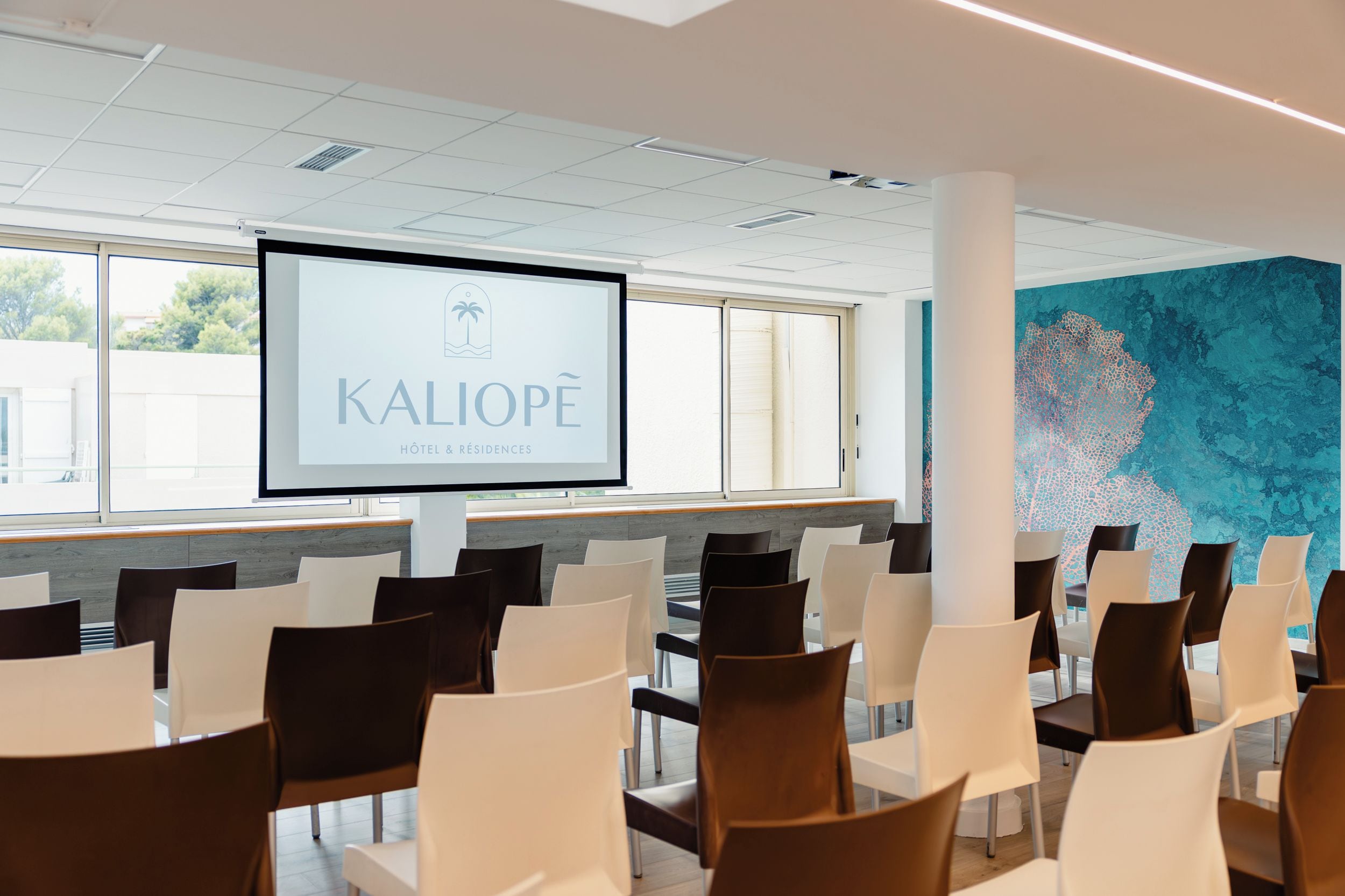 Une salle de conférence équipée d’un écran de présentation affichant le logo de « KALIOPE HOTEL & RESTANCES », avec des sièges disposés de manière ordonnée et une grande fresque abstraite sur le mur.