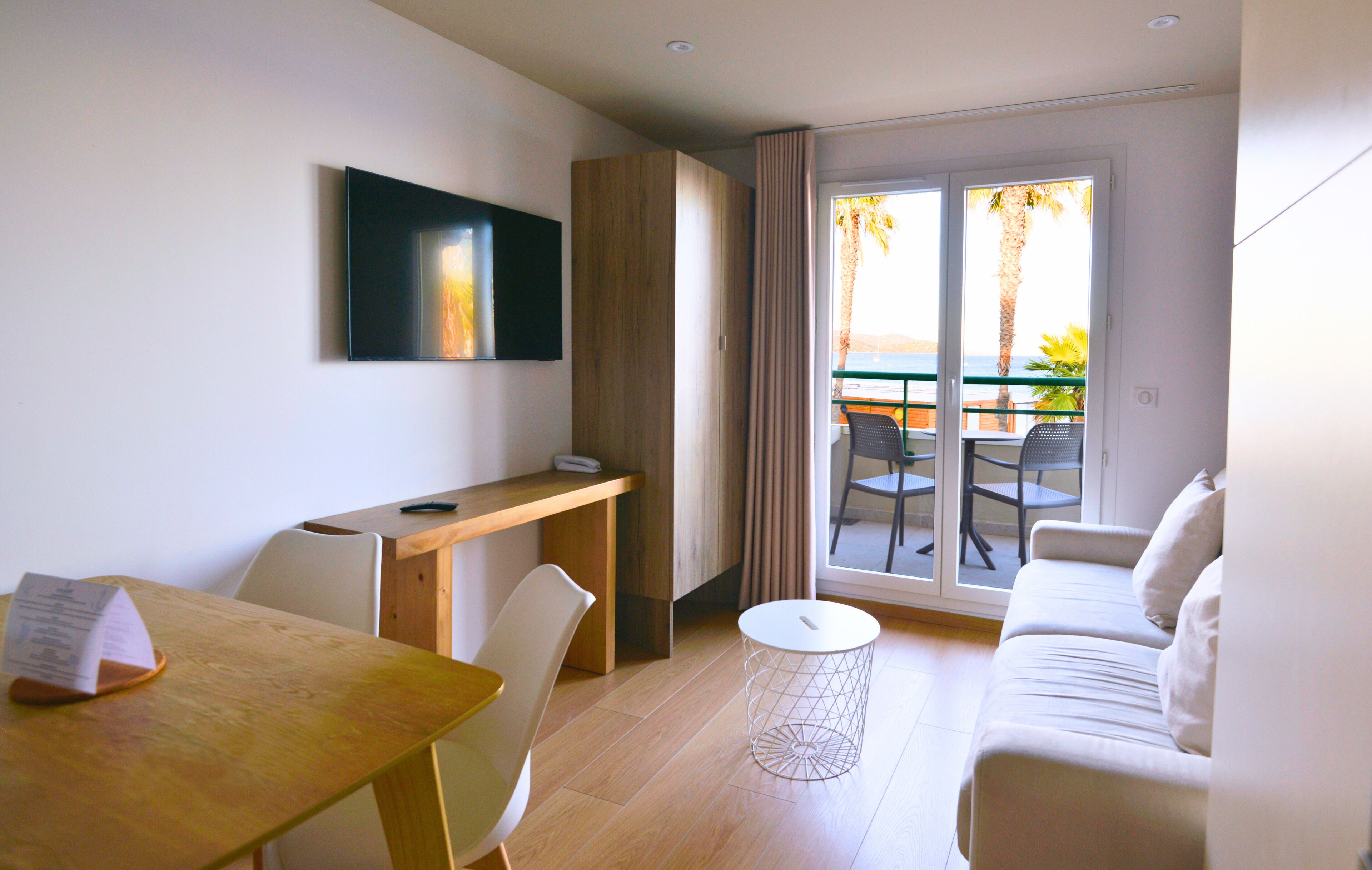A modern hotel room with a wooden desk, a white sofa, a dining table, and a TV mounted on the wall.