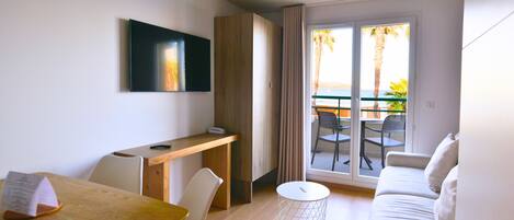 A modern hotel room with a wooden desk, a white sofa, a dining table, and a TV mounted on the wall.