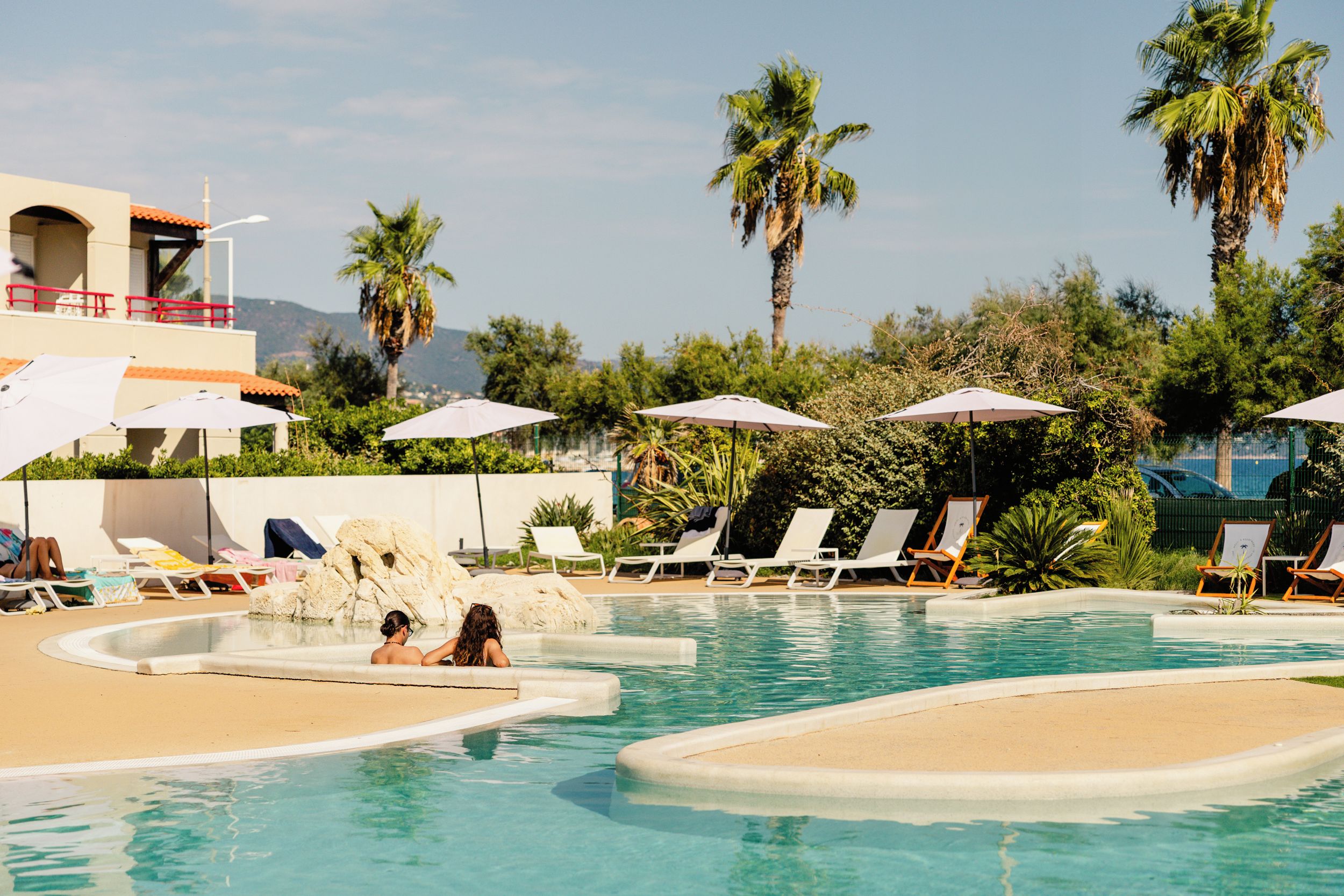 A pool area with lounge chairs and umbrellas, surrounded by palm trees and a building with a red railing.