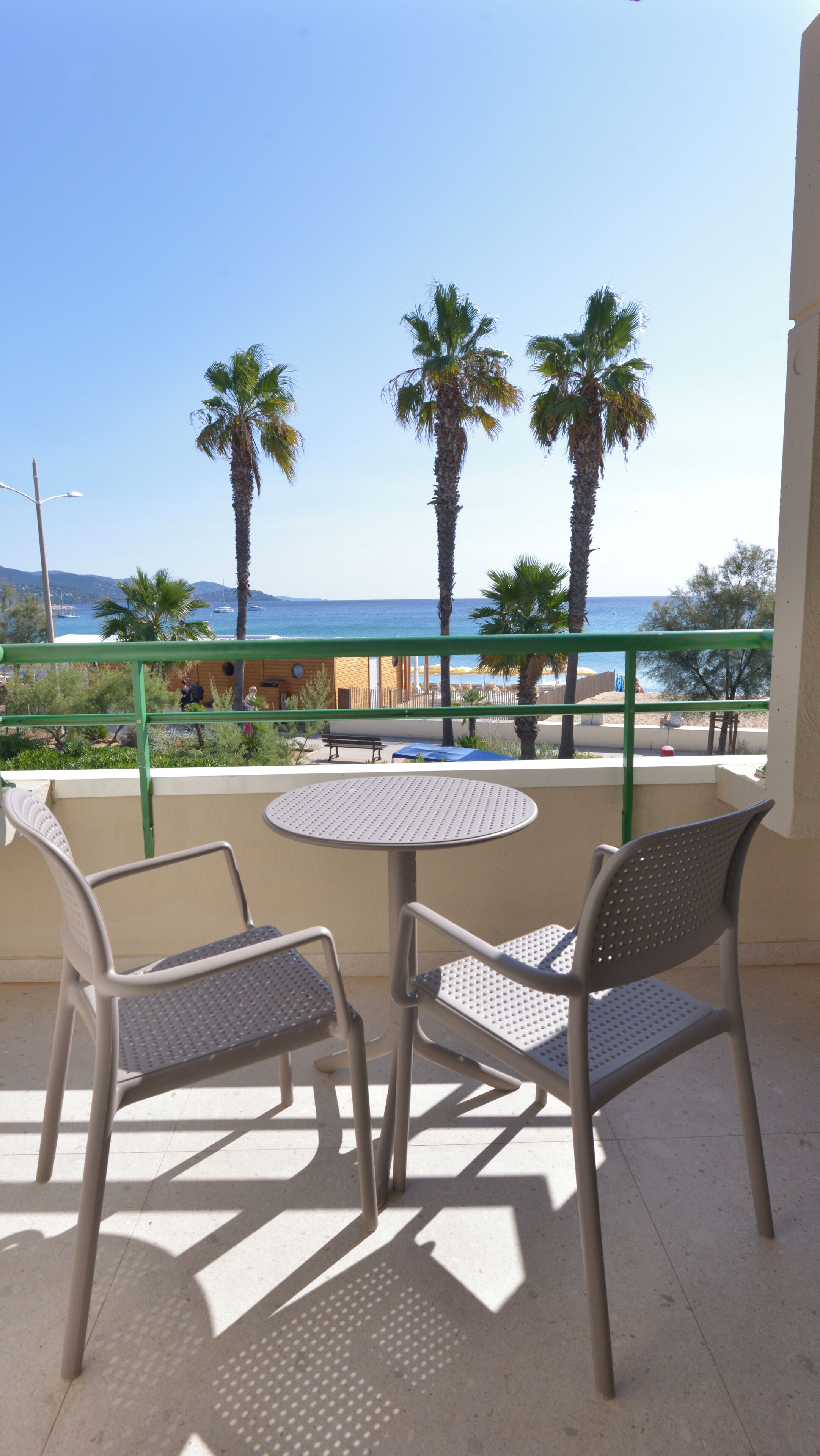 A balcony with two metal chairs and a small round table, overlooking a beach with palm trees and the sea.