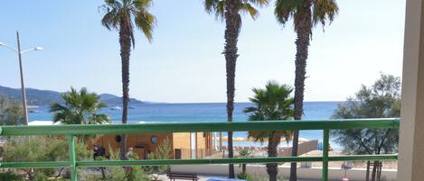 A balcony with two metal chairs and a small round table, overlooking a beach with palm trees and the sea.