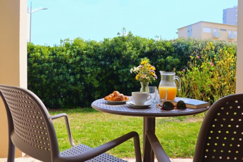 Une table ronde avec un croissant, une tasse, un pichet de jus d’orange et des lunettes de soleil, sur une terrasse ensoleillée agrémentée de chaises et d’un arbuste en arrière-plan.