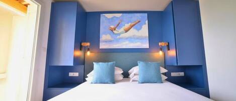 Ein Schlafzimmer mit einem großen Bett, zwei blauen Kissen, einem Bild von einer springenden Person und Wandleuchten.