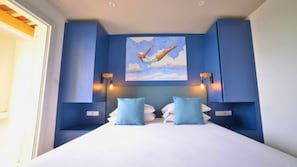A bedroom with a large bed, two blue pillows, a painting of a person jumping, and wall-mounted lights. - Kaliopé Hotel & Résidence by Oustal del Mar (Le Lavandou)