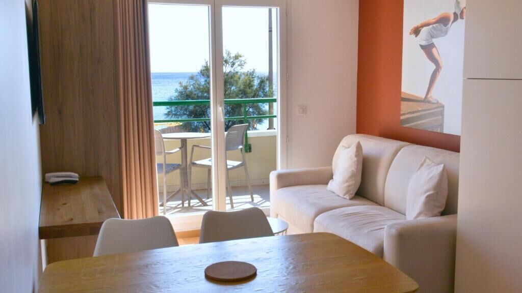 A living room with a sofa, a dining table, and a view of the sea through a glass door.