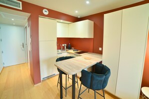 A compact kitchen with a wooden table and two blue chairs, a white refrigerator, and a white oven. - Kaliopé Hotel & Résidence by Oustal del Mar (Le Lavandou)