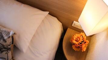A neatly made bed with a white pillow and a decorative pillow with palm tree designs. There is a small wooden nightstand with a white lamp and a bouquet of orange flowers.