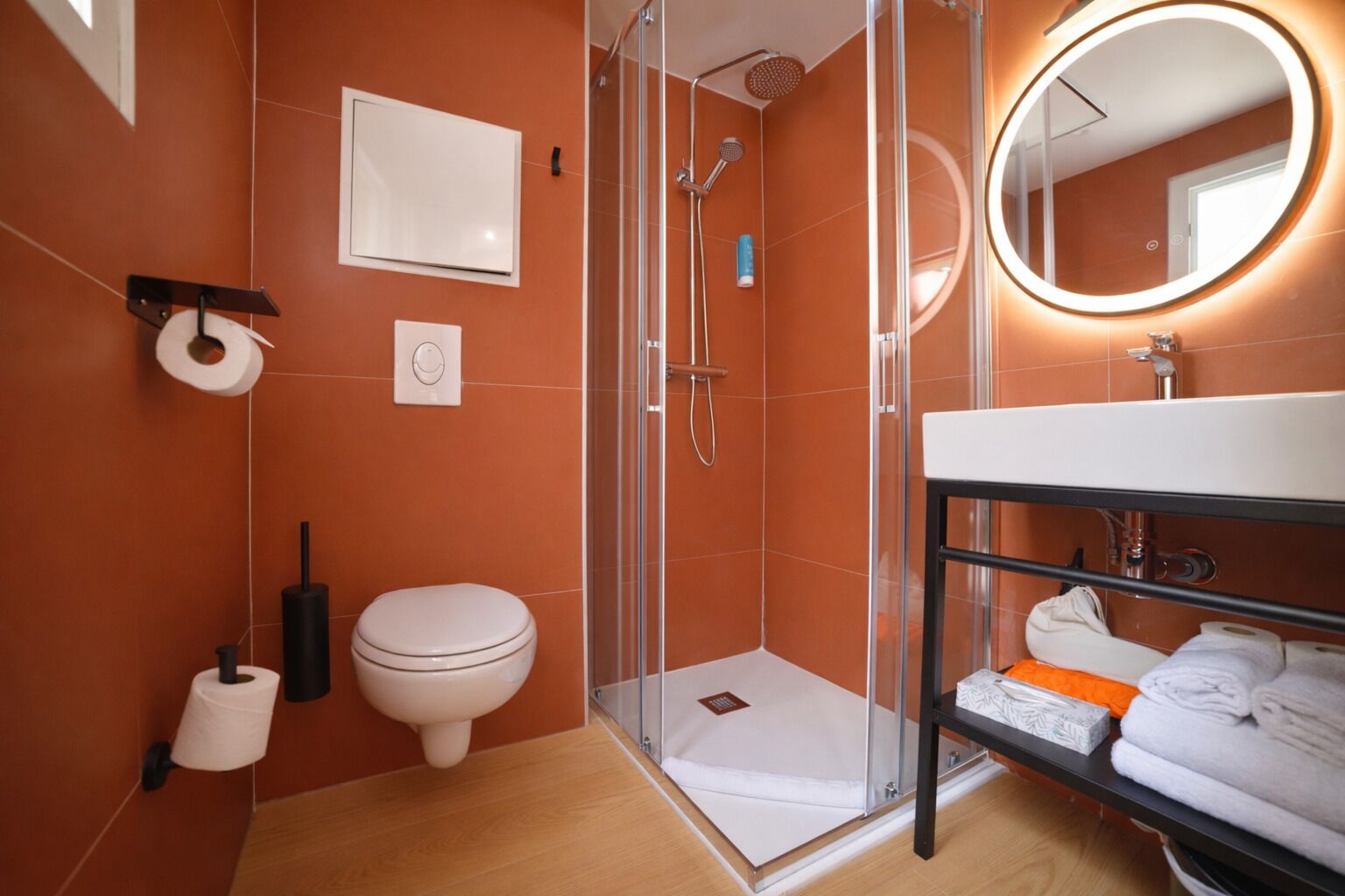 A modern bathroom with a glass-enclosed shower, a white toilet, a sink with a mirror, and a shelf with towels.