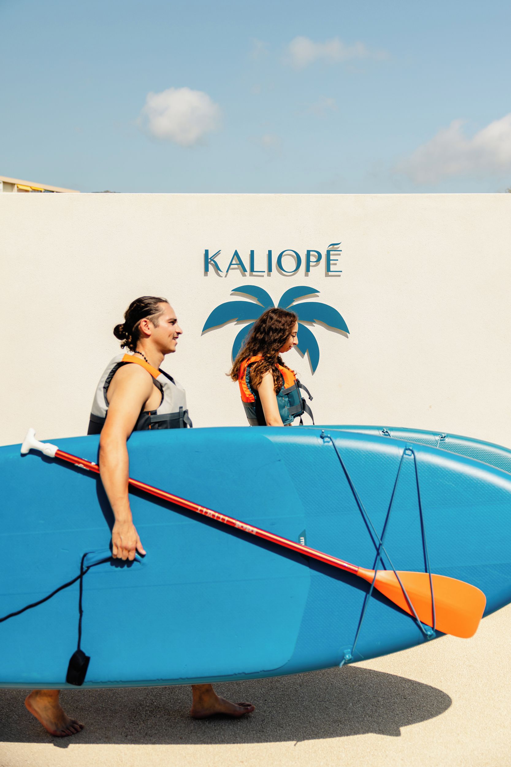 Zwei Personen stehen neben einem blauen Boot mit einem Palmen-Logo und dem Schriftzug 'KALIOPE' an einer Wand.