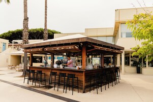 Un bar al aire libre con estructura de madera, taburetes de bar y una persona sirviendo.