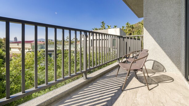 Un balcón con barandilla de metal, dos sillas de mimbre y vistas a un paisaje urbano con edificios y palmeras.