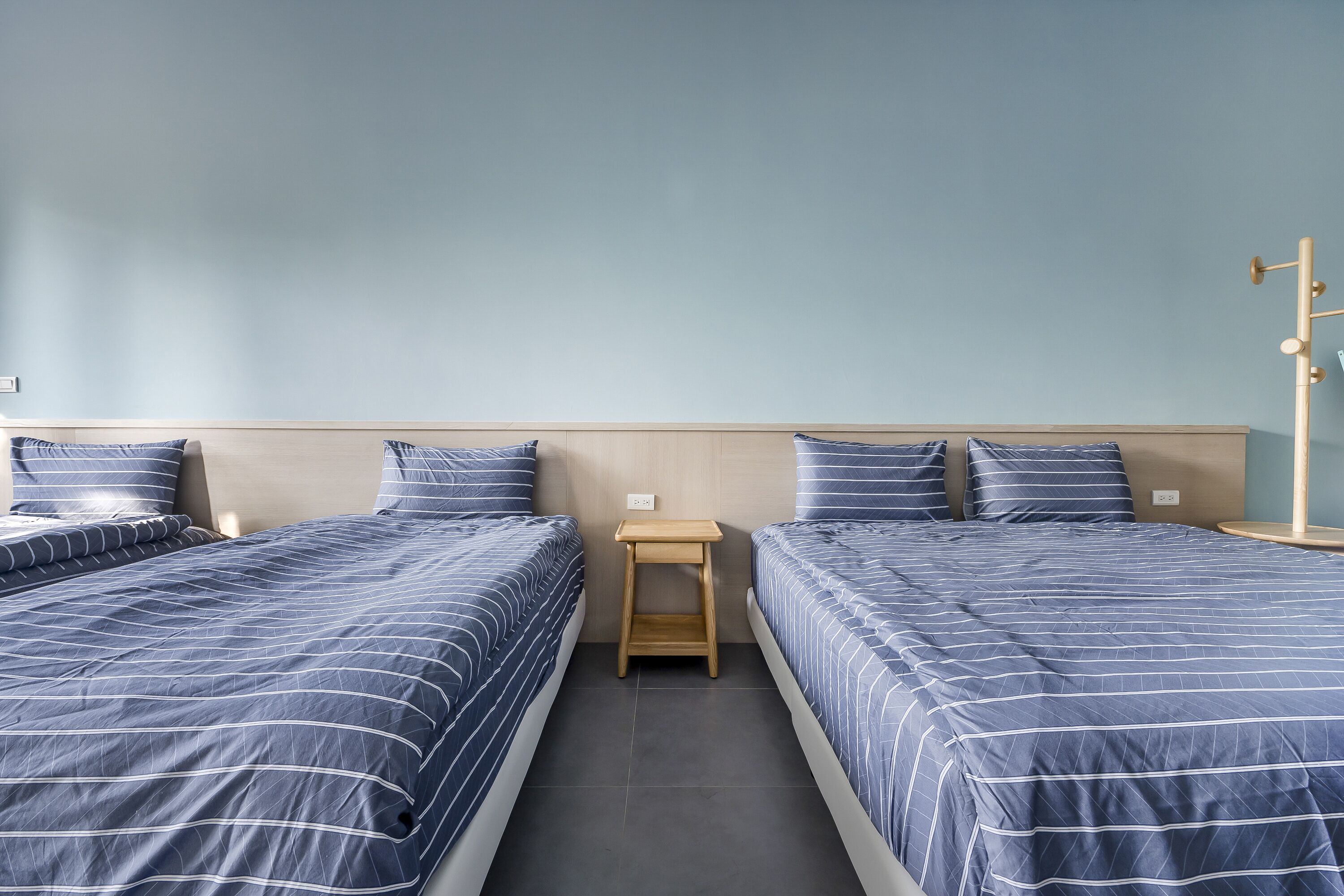 A room with two beds, each with blue bedding and white stripes, a small wooden nightstand, and a light blue wall.