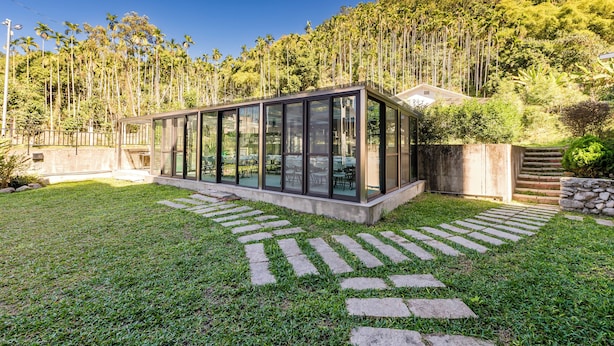 A modern glass-enclosed structure with a stone pathway leading to it, surrounded by a well-maintained lawn and tall trees. - Hills Valleys (Puli)