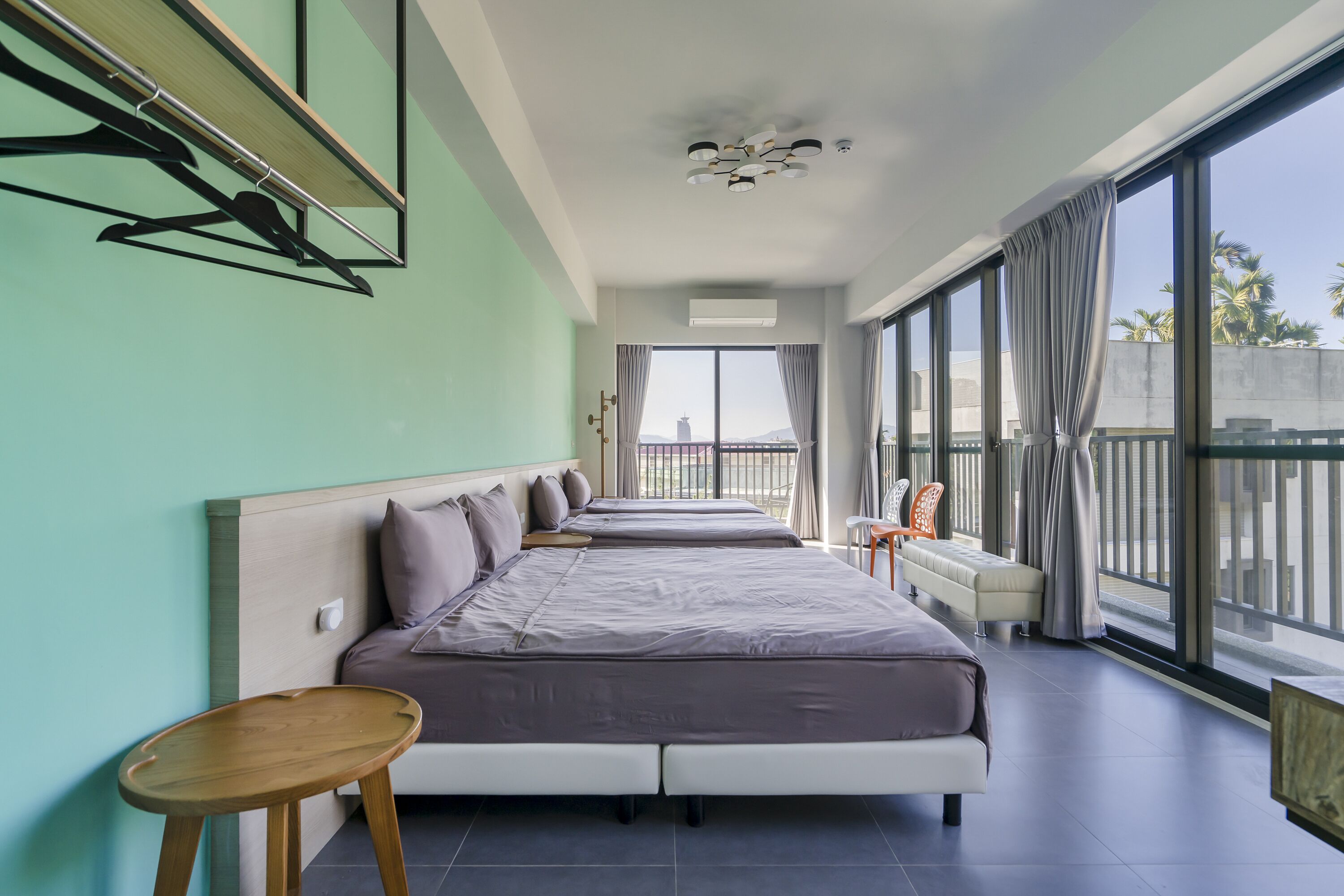 A modern hotel room with two beds, a small table, a chair, and large windows offering a view of the city.