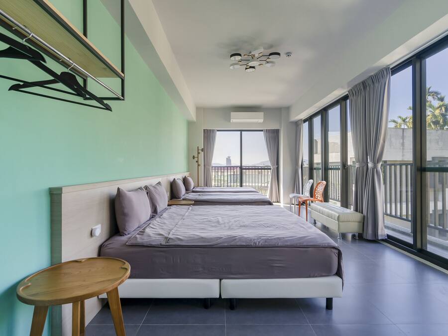 A modern hotel room with two beds, a small table, a chair, and large windows offering a view of the city.