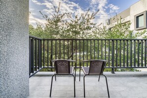A balcony with two wicker chairs, a metal railing, and a view of trees and buildings. - Hills Valleys (Puli)