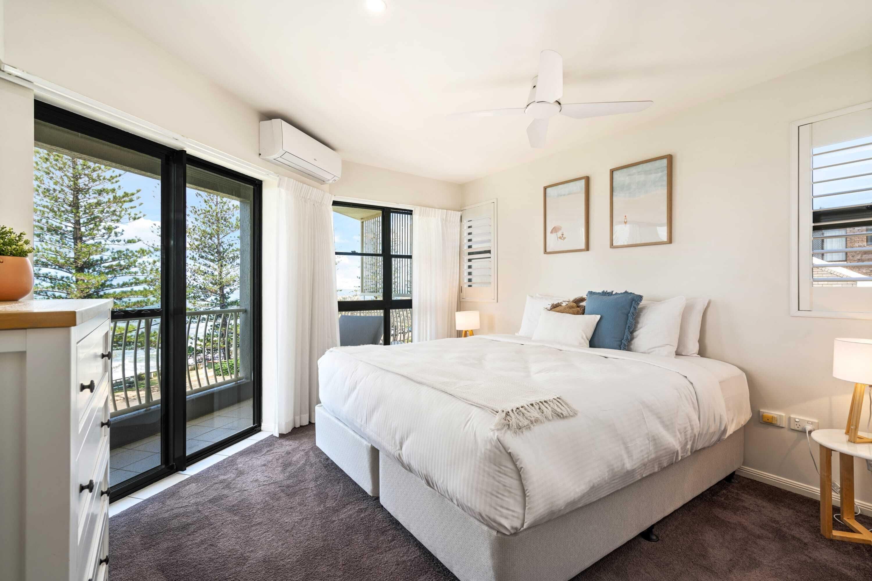 A bedroom with a large bed, a sliding glass door to a balcony, a ceiling fan, and a small table with a lamp.