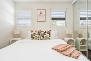 A bedroom with a bed, two bedside tables, a mirror, and a window with blinds.
