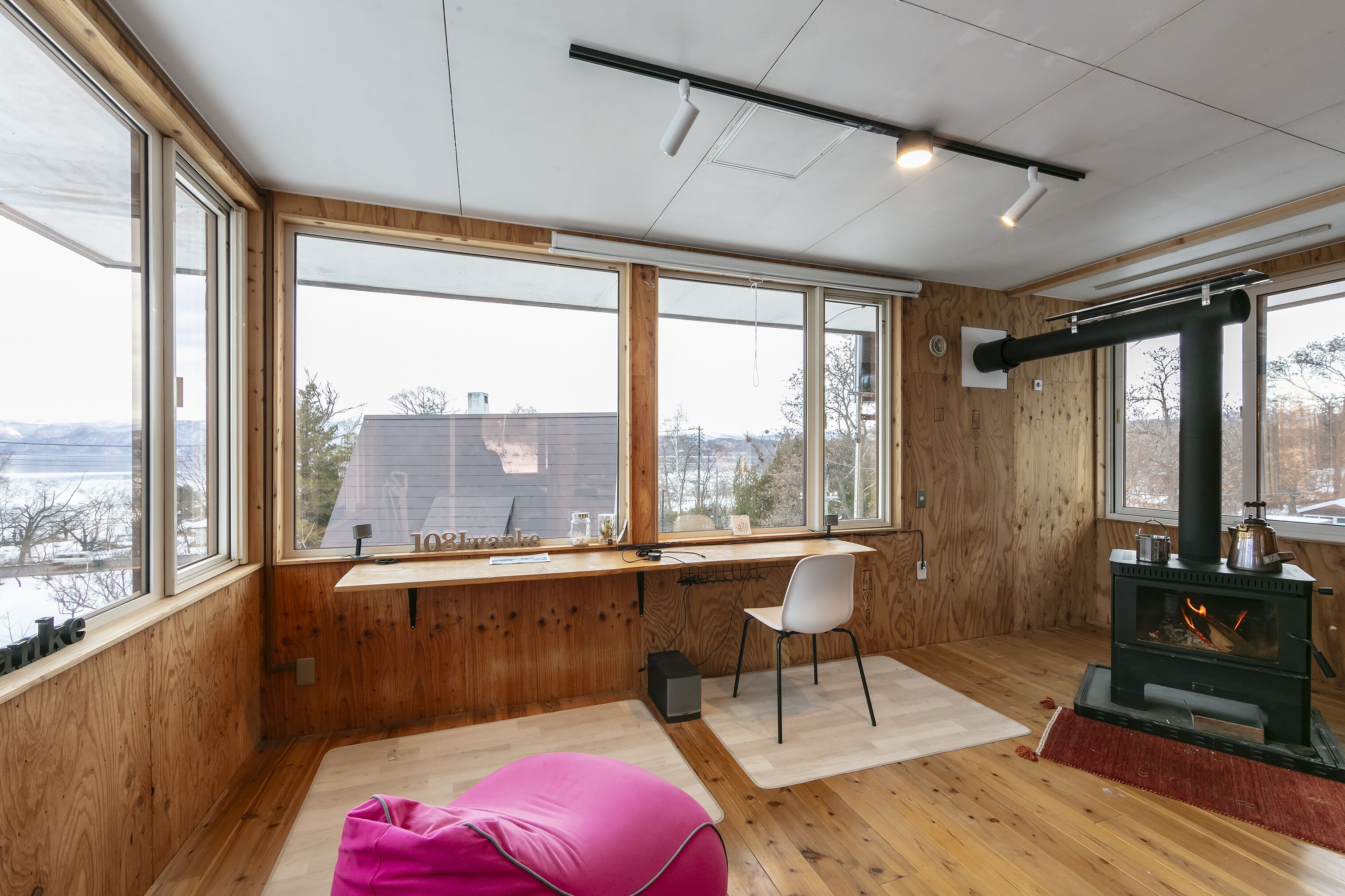 A cozy room with large windows, a wooden desk, a chair, a pink bean bag, and a wood-burning stove.