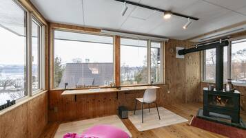 A cozy room with large windows, a wooden desk, a chair, a pink bean bag, and a wood-burning stove.