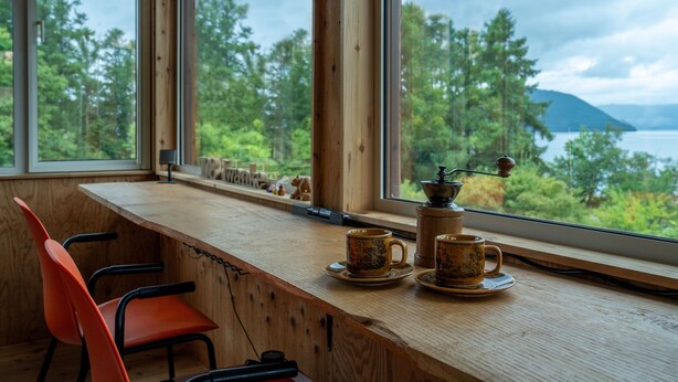 Un tavolo di legno con due tazze e piattini, una sedia rossa, e vista su una foresta e un lago attraverso ampie finestre.