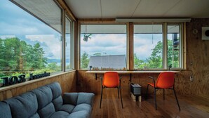 Una stanza con pavimento in legno, un divano grigio, due poltrone arancioni e ampie finestre che si affacciano su alberi e una casa.