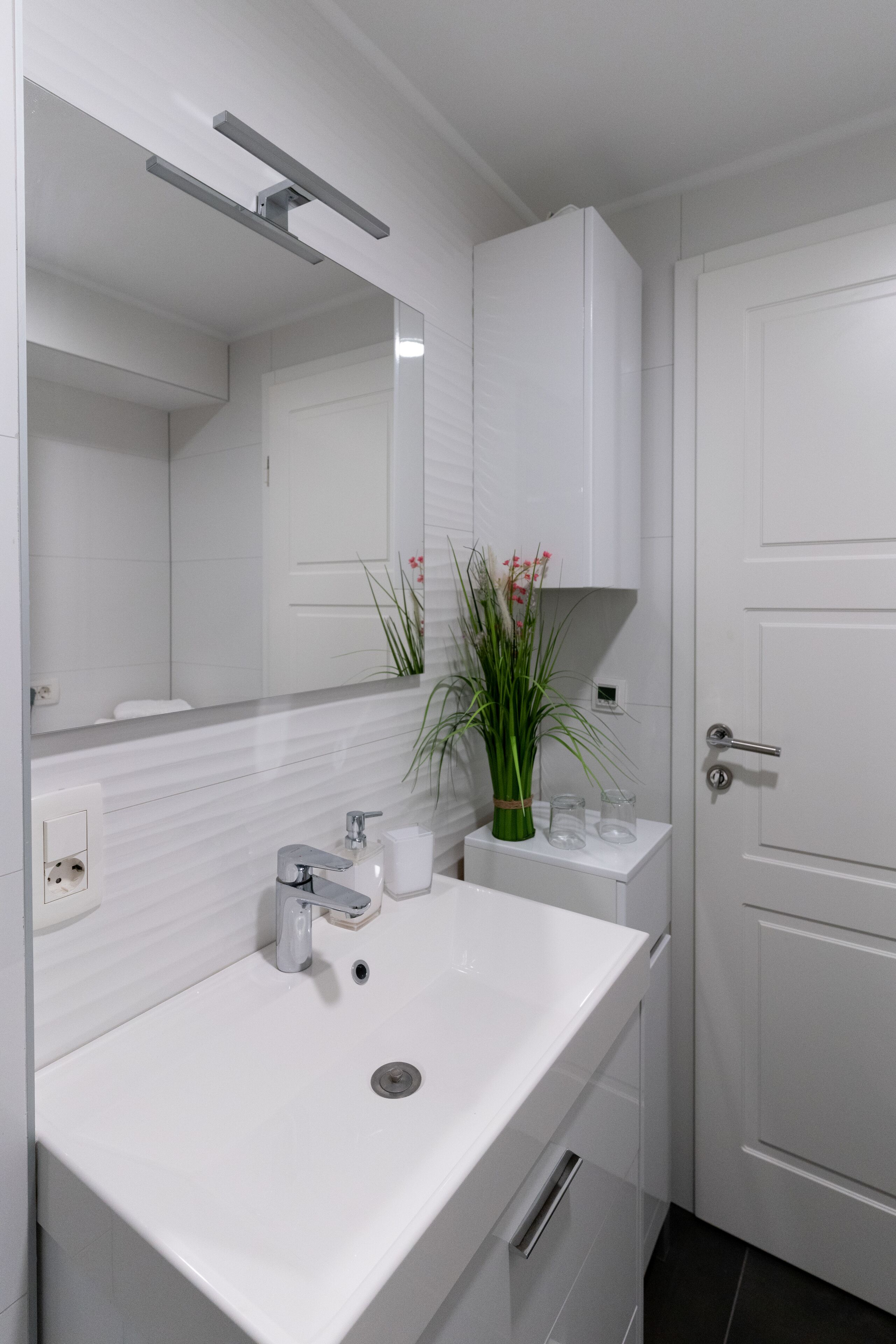 A modern bathroom with a white sink, mirror, and cabinets. There is a plant on the counter and a door to the right.
