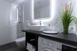 A modern bathroom with a white sink, mirror, and toilet. There is a shower area with a glass door and a washing machine in the corner. - Bonne Chance Apartments (Zagreb)